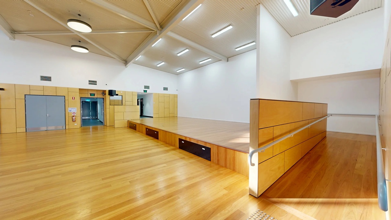 A spacious room with a wooden floor and a high ceiling featuring exposed beams. On the left, there are light-coloured wooden panels with a grey double door and an emergency exit. In the centre, a raised wooden stage with a ramp and handrail extends into the room. The right side has a continuation of the stage and a white wall. The ceiling has circular and rectangular lights, providing ample illumination.
