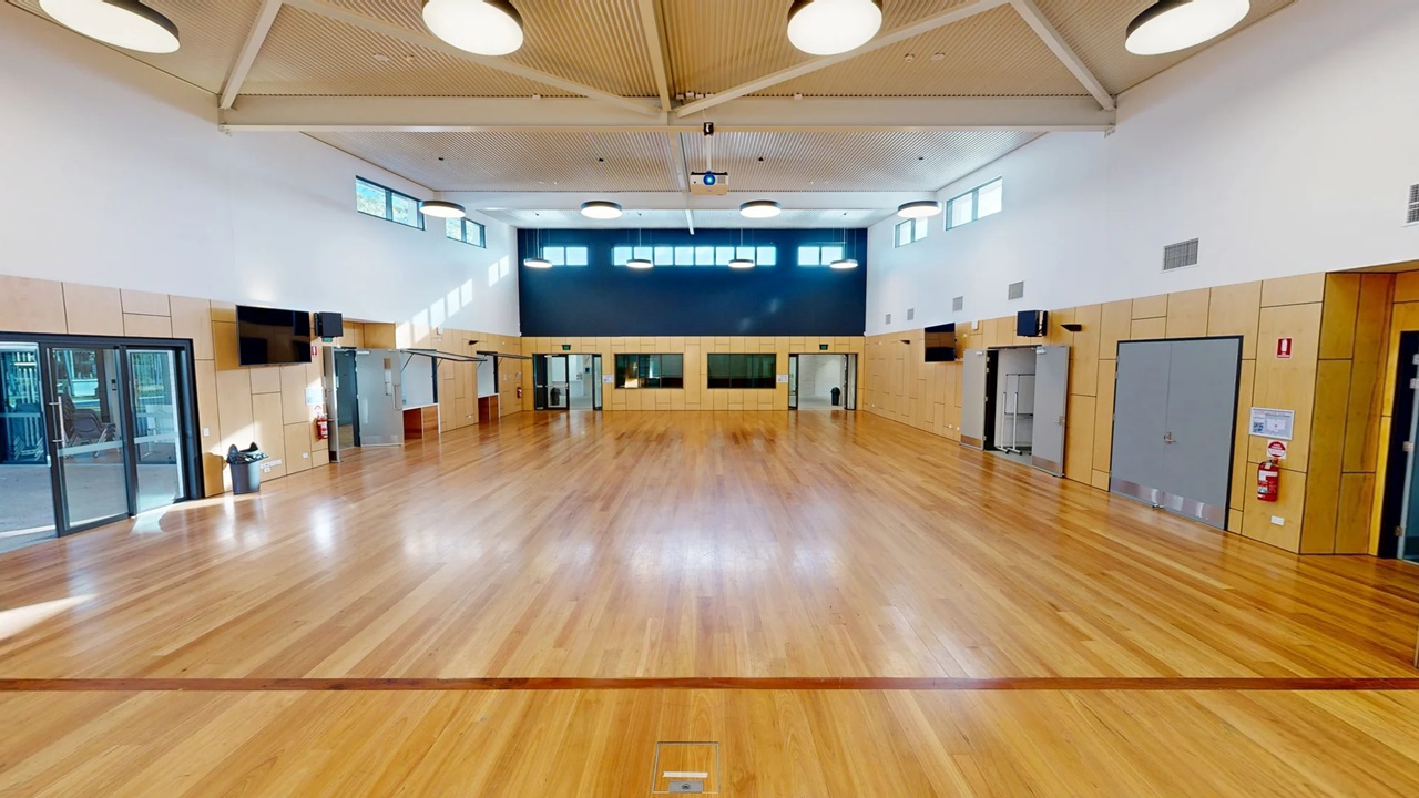 A spacious hall with a polished wooden floor and high ceiling featuring circular lights. The walls are lined with light-coloured panels, and there are several doors and windows along the sides. At the far end, a dark blue accent wall with high windows adds contrast. A projector is mounted on the ceiling, and there are fire extinguishers and a few wall-mounted screens visible. The room is well-lit with natural light streaming in from the windows.