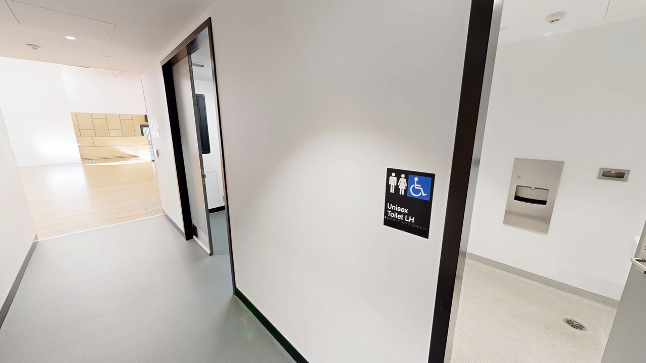 A corridor with light grey flooring and white walls. On the right, a door is slightly open, revealing a unisex toilet sign with male, female, and wheelchair symbols. Inside the room, a paper towel dispenser is visible on the wall. The ceiling is white with recessed lighting. In the background, there is a bright area with wooden cabinets and a counter.