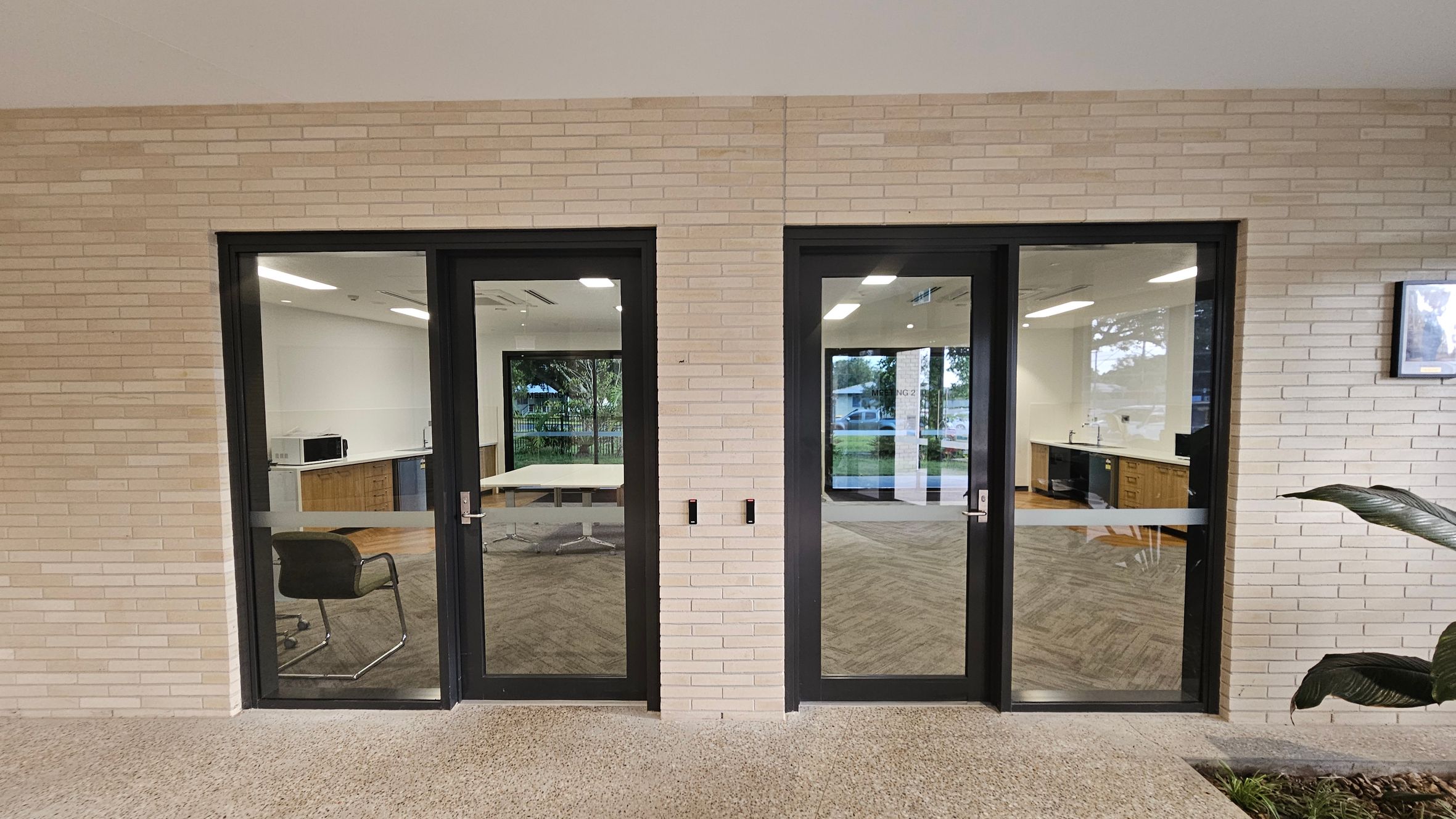 Two glass doors with black frames are set in a light brick wall. The left door reveals a room with a chair and a table, while the right door shows a kitchenette with wooden cabinets and a sink. The floor is speckled concrete, and a large plant is partially visible on the right. Reflections in the glass show outdoor greenery and a parked car.