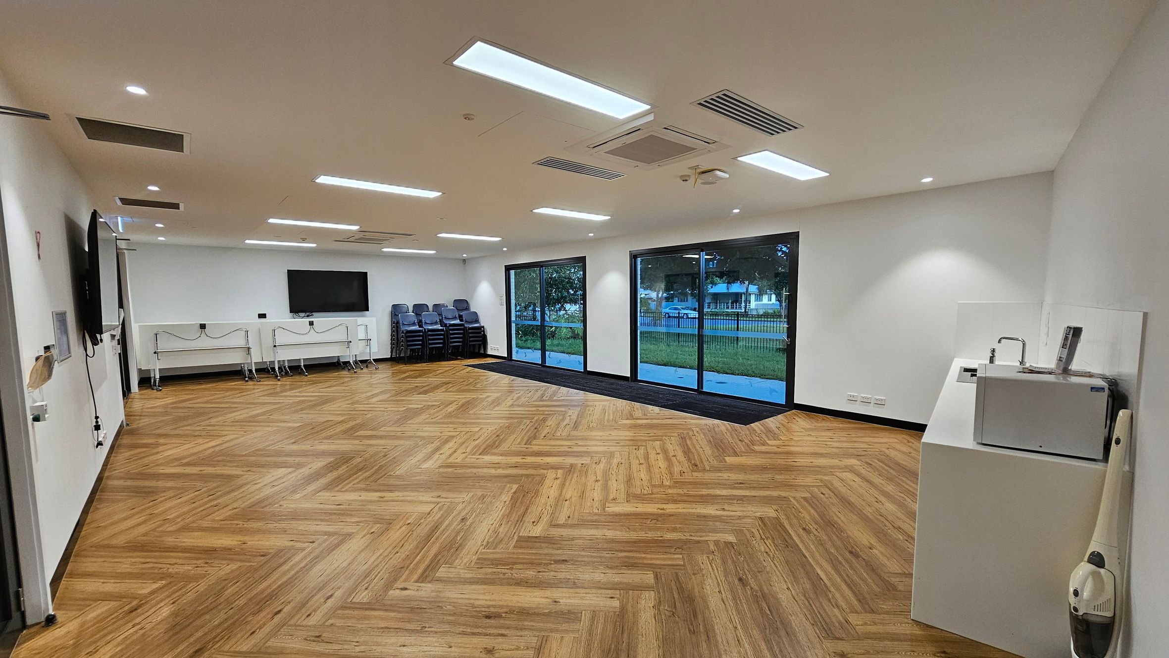 A spacious room with a herringbone-patterned wooden floor. On the left, there are two wall-mounted screens and several stacked chairs. The back wall features large glass sliding doors, offering a view of a grassy area outside. On the right, there's a water cooler and a vacuum cleaner. The ceiling is equipped with recessed lighting and air conditioning vents.