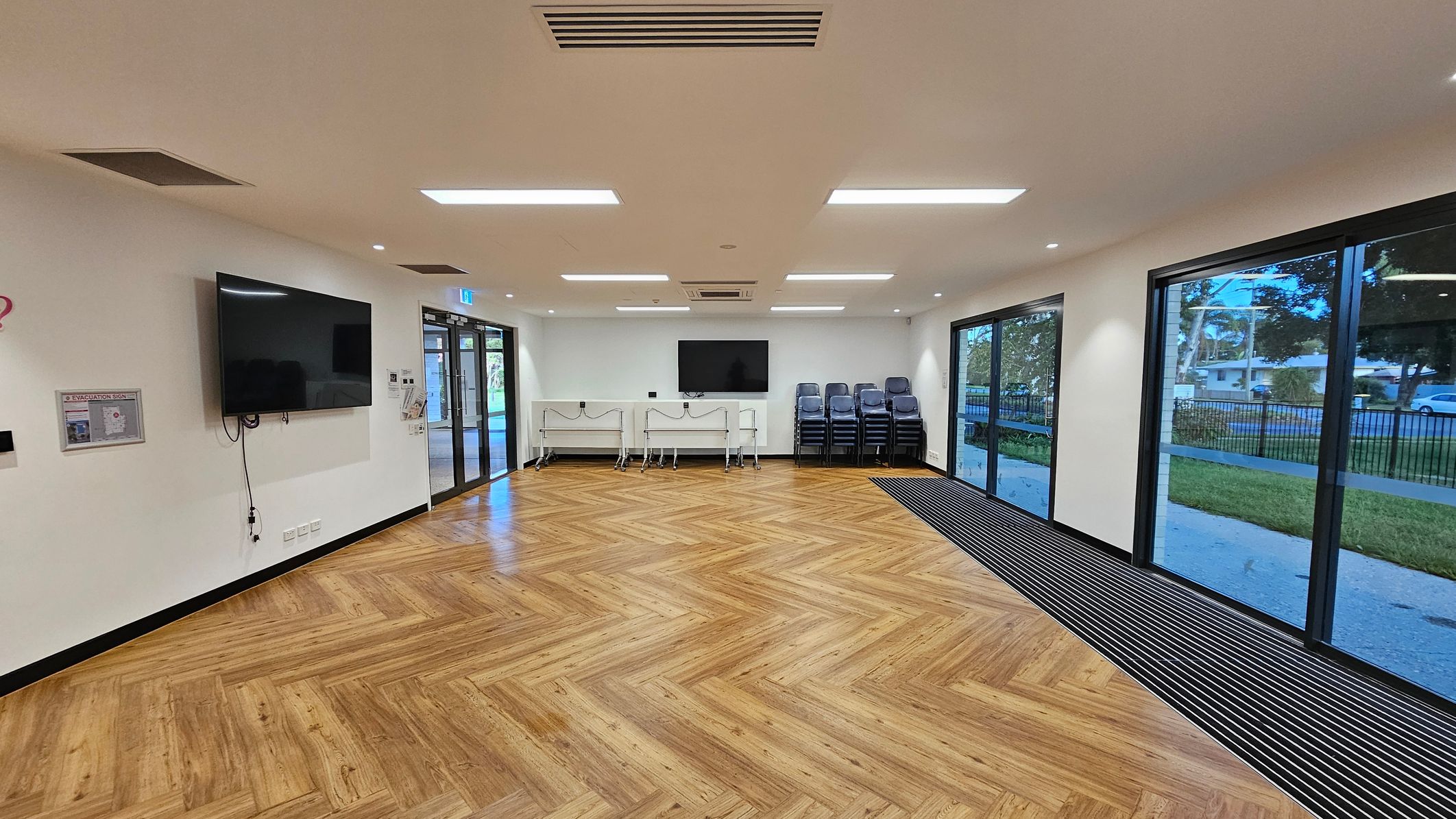 A spacious room with a wooden herringbone-patterned floor. On the left wall, a large TV is mounted above an evacuation plan. The back wall features another TV, two foldable tables, and a stack of chairs. The right side has large glass doors and windows, offering a view of a grassy area and a street outside. The ceiling is fitted with rectangular lights and air vents.