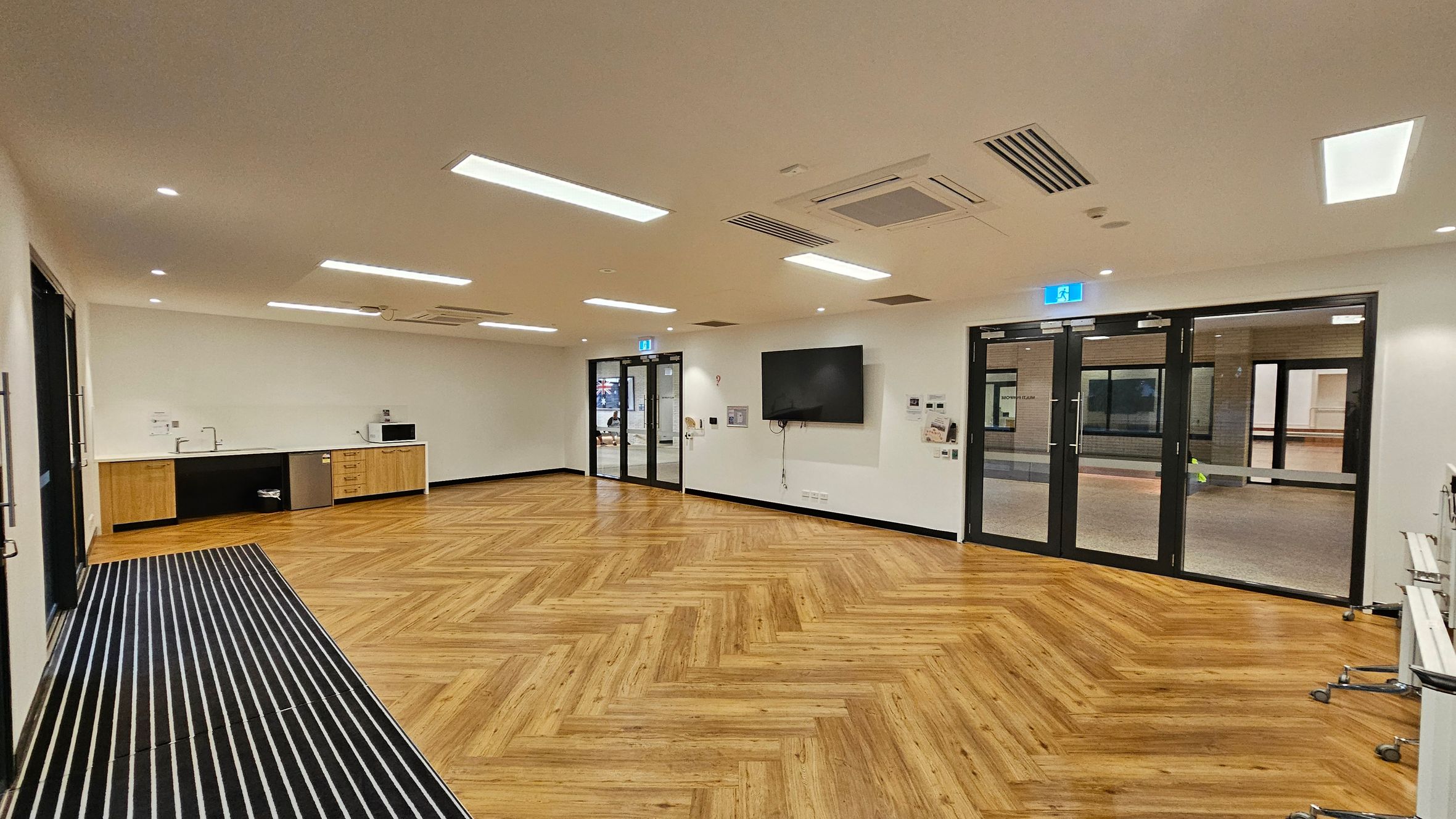 A spacious room with a herringbone-patterned wooden floor. On the left, there is a kitchenette with a sink, microwave, and small fridge. The centre features a wall-mounted television and a set of glass doors with an exit sign above. On the right, there are more glass doors leading to another area. The ceiling has recessed lighting and air conditioning vents. A striped mat is on the floor near the entrance.