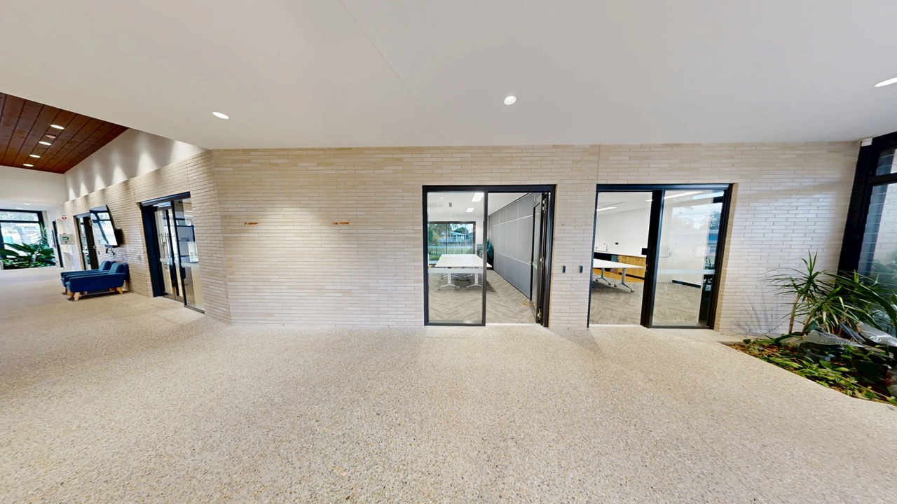 A modern interior space with a light-coloured, textured wall featuring two glass doors. The left door opens to a room with a table and chairs, while the right door reveals a room with yellow chairs. On the left, a blue sofa is placed near a window with plants visible outside. The floor is carpeted in a neutral tone, and the ceiling is white with recessed lighting. On the right, there is a small indoor garden with green plants.