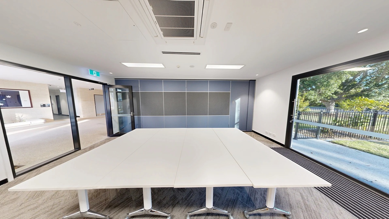 A modern conference room with a large white table in the centre on a wooden floor. The left side features glass doors leading to a hallway with visible exit signs. The right side has large windows showing a view of trees and a fenced outdoor area. The back wall is covered with grey panels. The ceiling is white with recessed lighting and an air conditioning vent.