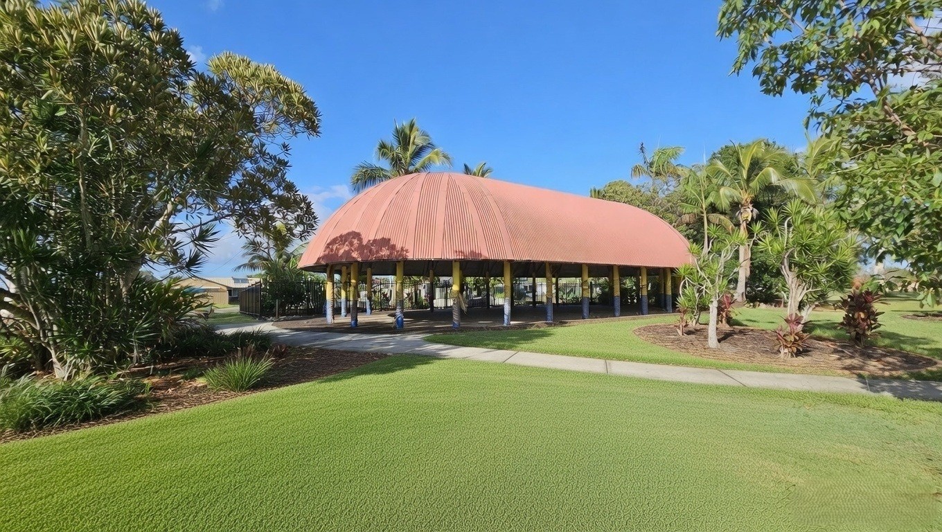 A large, open-sided structure with a curved, terracotta-coloured roof supported by yellow pillars stands in the centre. Surrounding the structure are lush green lawns and various trees, including palm trees and other tropical plants. A paved pathway leads towards the building, and the sky is clear and blue.