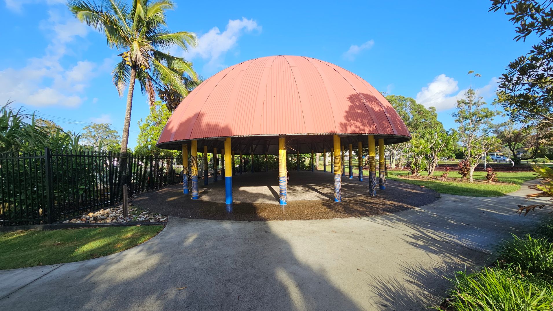 A large, dome-shaped structure with a red roof is supported by yellow and blue pillars. The ground is a mix of concrete and grass. On the left, a tall palm tree stands next to a black metal fence with lush green plants behind it. On the right, there are more trees and a parked car in the distance. The sky is clear and blue, casting shadows on the ground.