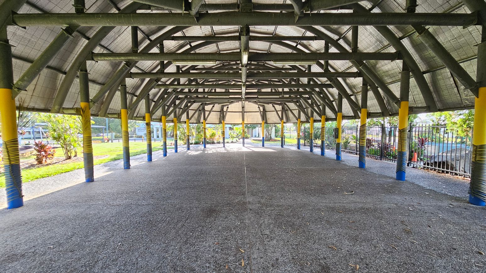 A large, open-sided structure with a high, arched roof supported by numerous green and yellow pillars. The floor is grey and textured. On the left and right, there are metal fences and greenery, including small trees and shrubs. In the background, more plants and a glimpse of a building are visible. The ceiling is made of a grid-like material, allowing light to filter through.