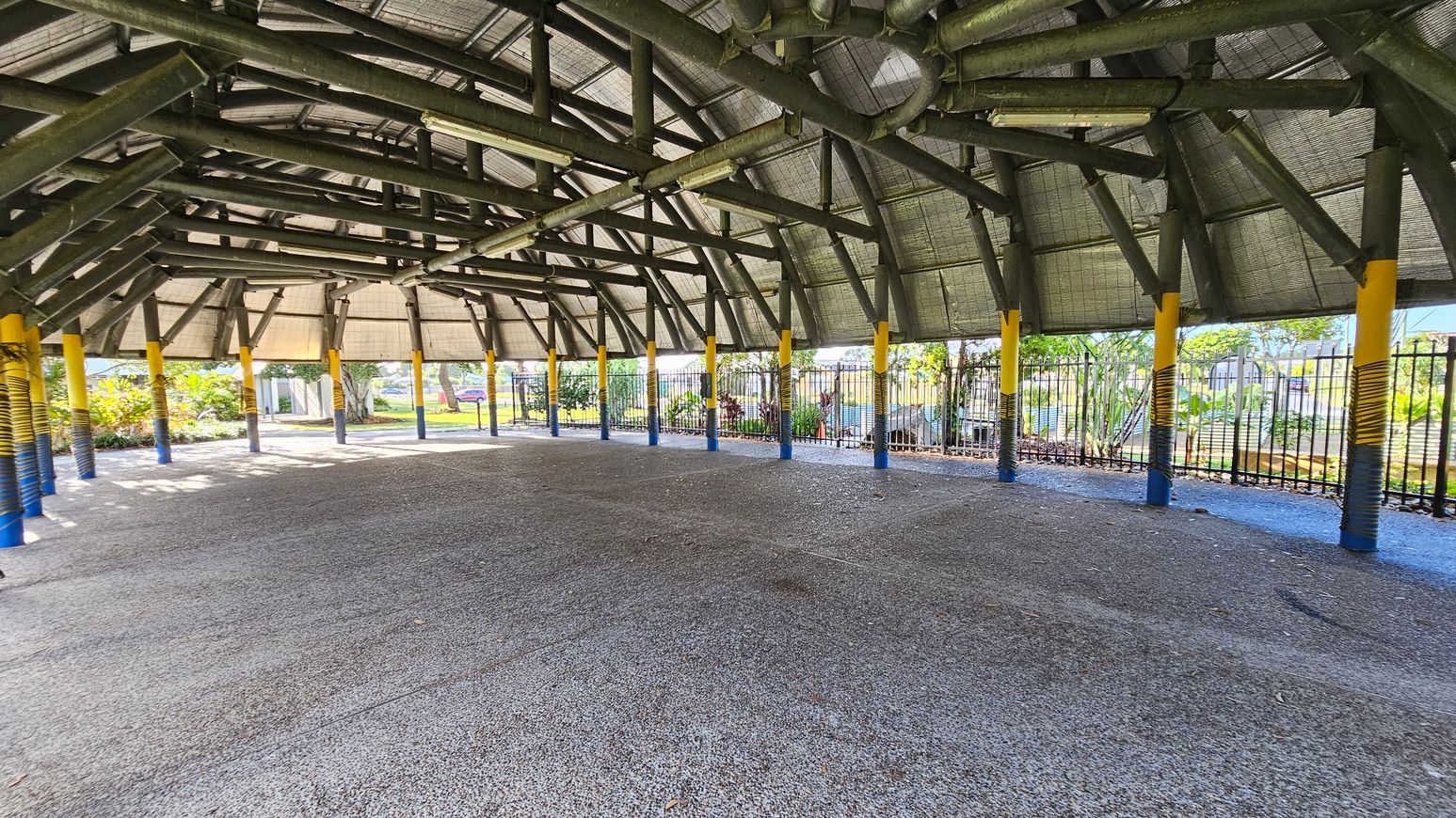 A large open pavilion with a high, thatched roof supported by numerous yellow and blue painted pillars. The floor is made of textured concrete. In the background, there is a metal fence with greenery and trees visible beyond it. The structure is well-lit by natural light, and the ceiling features exposed beams and lighting fixtures.