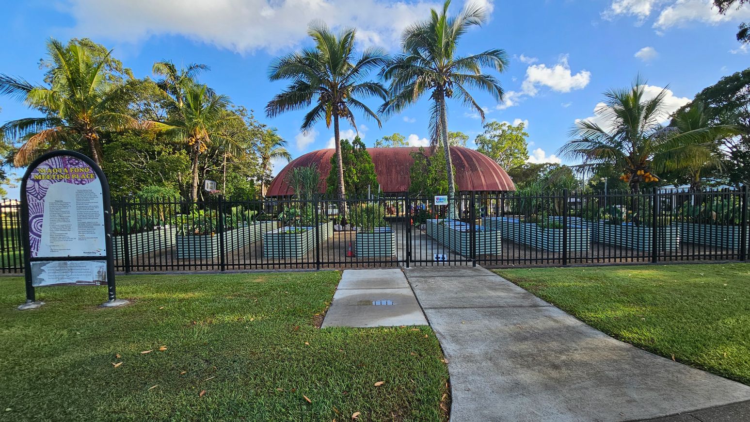 A garden area with a domed, rust-coloured building in the centre, surrounded by palm trees. In the foreground, a black metal fence encloses the space, with raised garden beds visible inside. A sign on the left reads "Maota Fono Meeting Place" with text beneath it. The ground is covered with grass and a concrete path leads up to the fence. The sky is blue with scattered clouds.