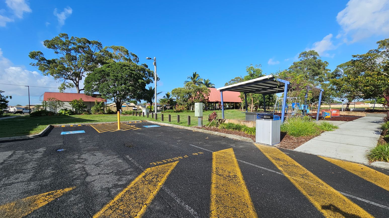 A car park with yellow directional arrows and a disabled parking space marked on the asphalt. On the right, a covered area with a bin and a pathway leading to a playground with swings. On the left, a grassy area with large trees and a building with a red roof. In the background, more trees and a clear blue sky.