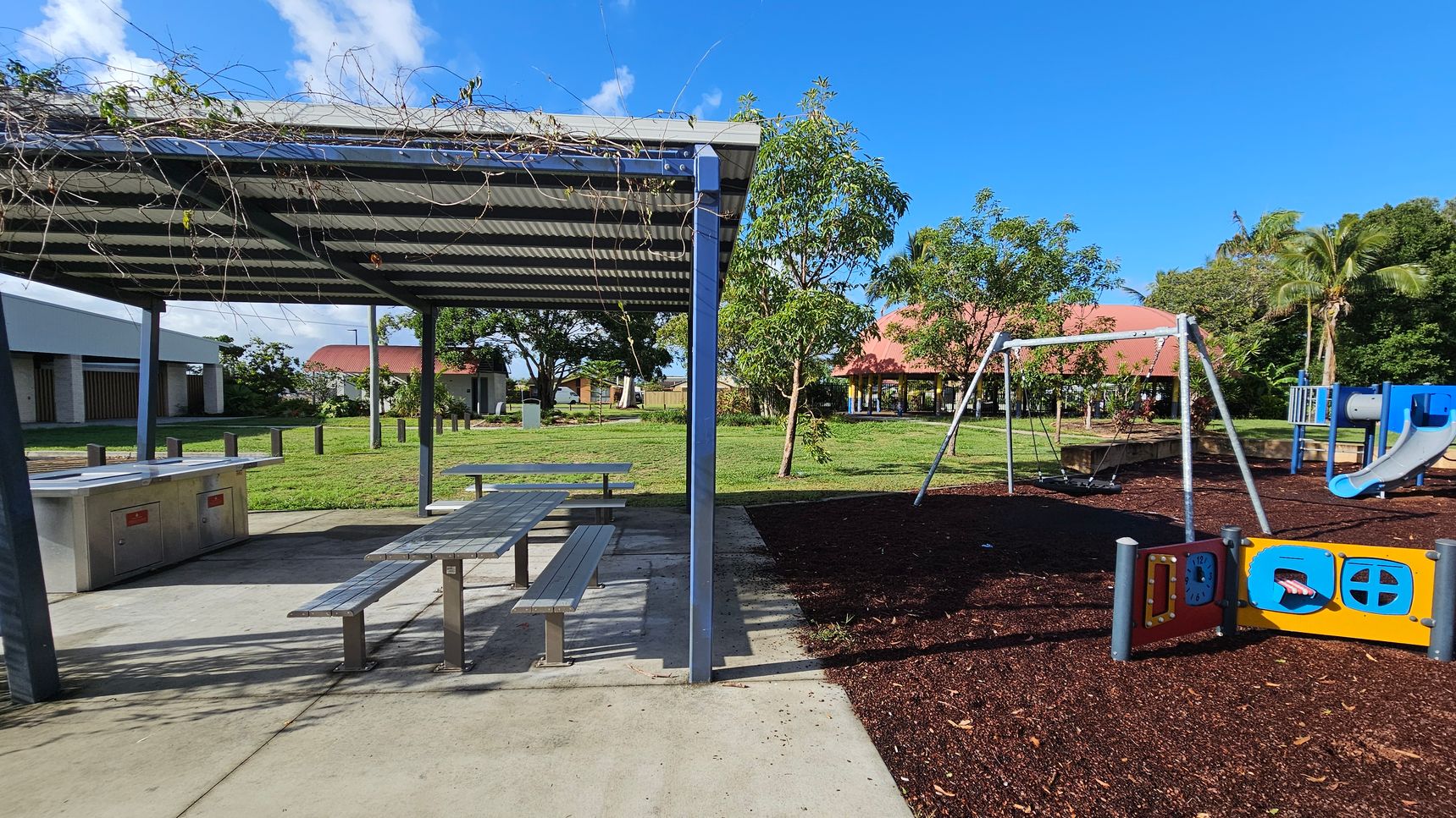 A park area with a covered picnic spot on the left, featuring a metal roof and a picnic table beneath. Nearby, there are barbecue facilities. On the right, a playground with a swing set and a blue slide is visible, set on a mulch surface. In the background, there are trees and a building with a red roof. The ground is a mix of concrete and grass, and the sky is clear.
