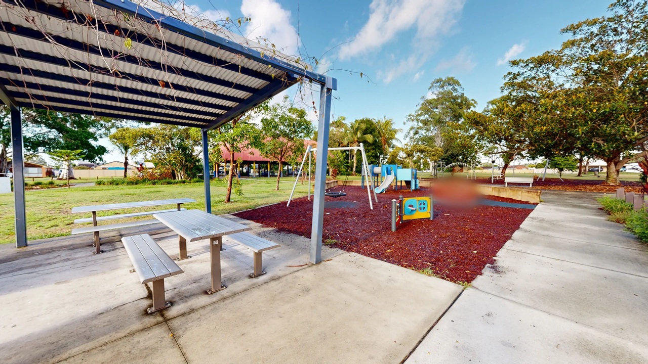 A playground with a covered picnic area on the left, featuring a metal table and benches on a concrete surface. To the right, there's a play area with swings, a slide, and a small climbing structure on a mulch surface. Trees and grass surround the area, with a pathway running alongside. In the background, there are more trees and a building with a red roof.