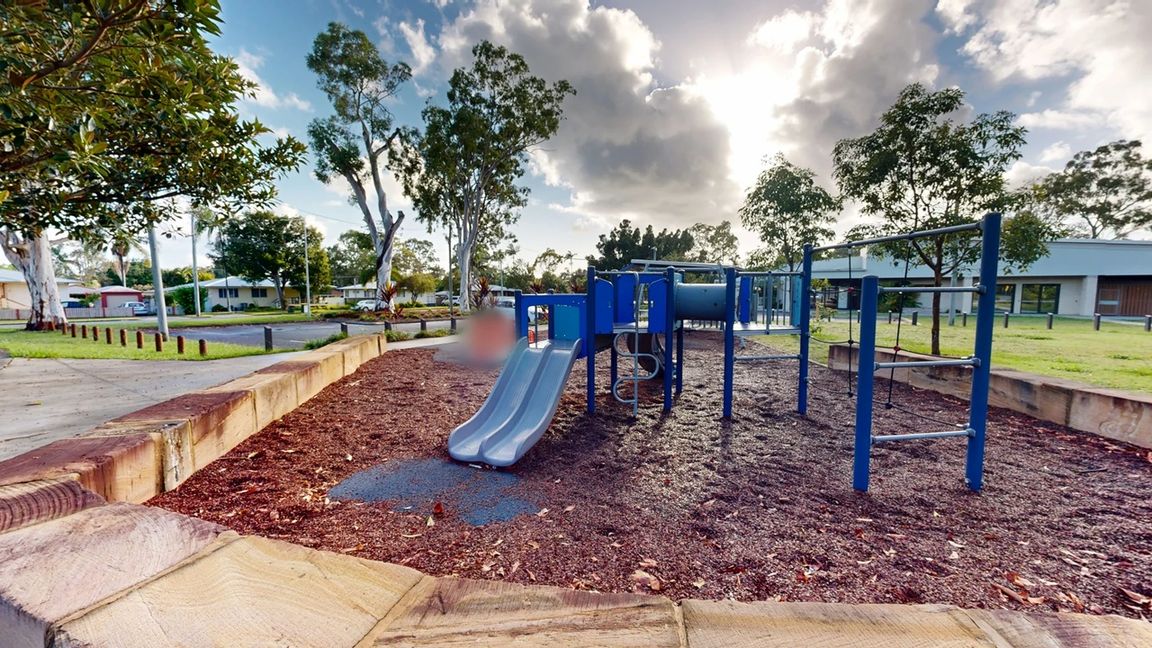A playground with a blue slide and climbing structure is in the centre, surrounded by a mulch-covered ground. On the right, there are additional climbing bars. In the background, there are trees and a building with a grassy area. To the left, a paved path and a row of bollards are visible, with more trees and residential houses in the distance. The sky is partly cloudy.
