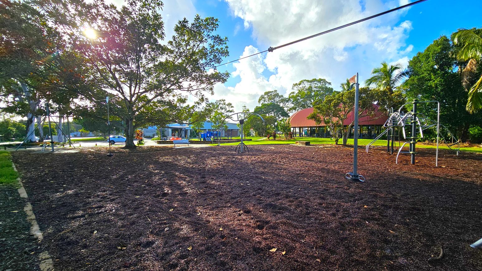 A playground with a mulch-covered ground. On the left, a large tree with sunlight filtering through its branches. In the centre, a zip line structure and a small roundabout. On the right, a climbing frame with metal bars. In the background, a building with a red roof and several trees, including palm trees. A road with a parked car is visible on the far left.
