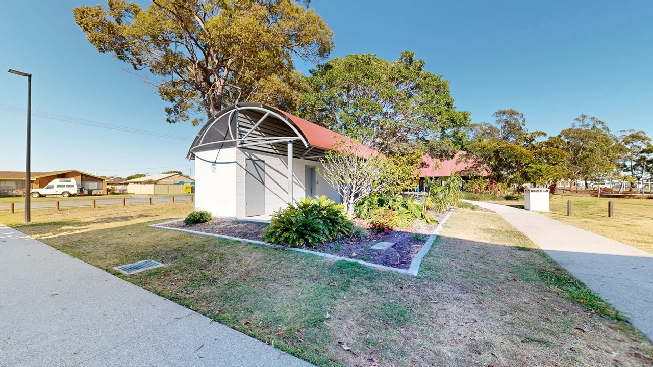 A small building with a curved red roof is in the centre, surrounded by a garden with various plants and shrubs. To the left, there's a paved path and a street with parked vehicles, including a white van. On the right, a concrete path leads through a grassy area with scattered trees. In the background, more trees and a clear blue sky are visible. A streetlamp is on the left side near the path.