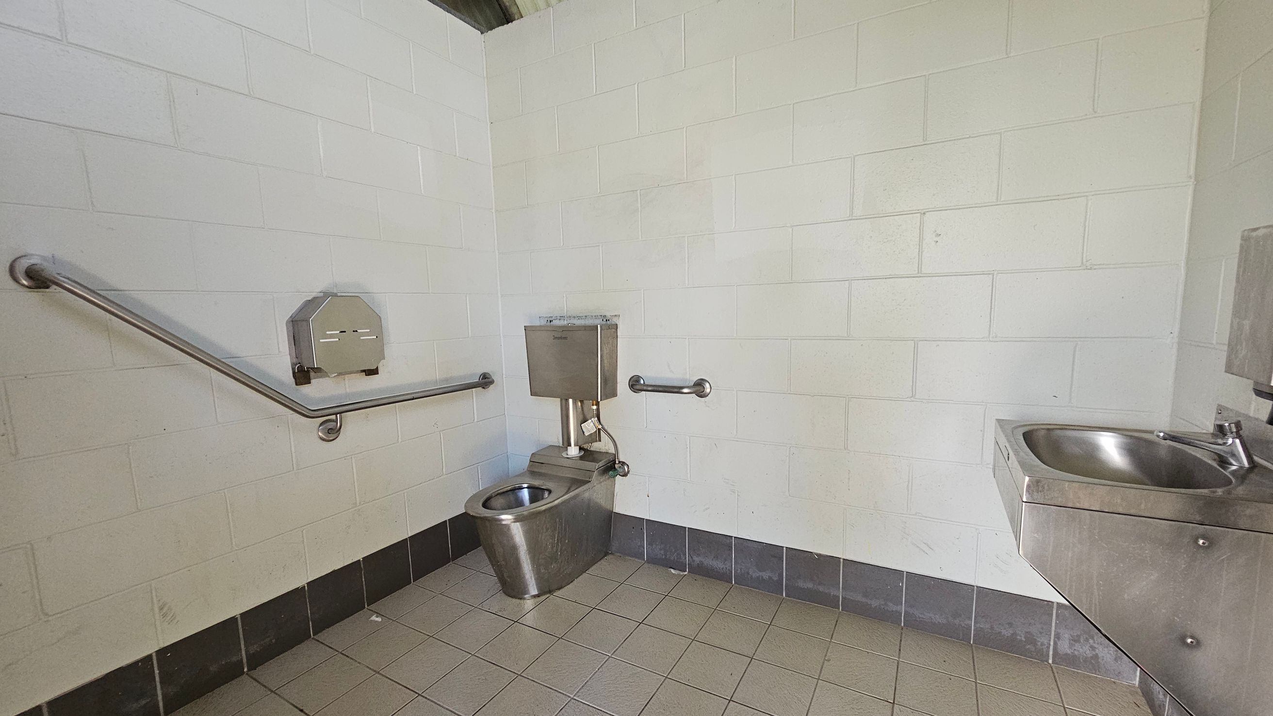A public restroom with white tiled walls and a tiled floor. On the left, there is a stainless steel toilet with a metal flush tank above it. A horizontal grab bar is mounted on the wall beside the toilet. On the right, a stainless steel sink is attached to the wall, with a soap dispenser above it. The floor is a mix of beige and grey tiles, and the ceiling is not visible.