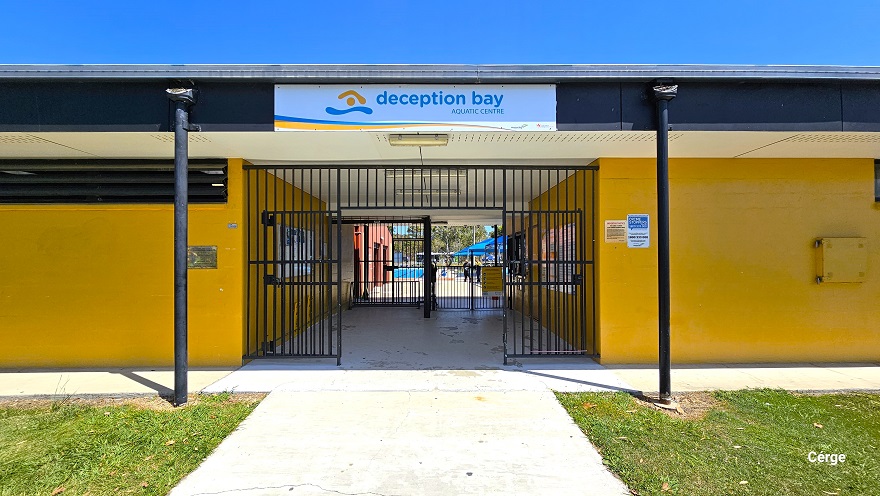 Concrete pathway with a rough-looking surface when walking from the right side of the main entrance of the brick mustard yellow corrugated iron building of Deception Bay Swimming Pool. The outdoor aquatic amenities can be seen through the chain-linked fence enclosing the property. Guests can pass through a silver payphone stand booth with a red logo at the top.