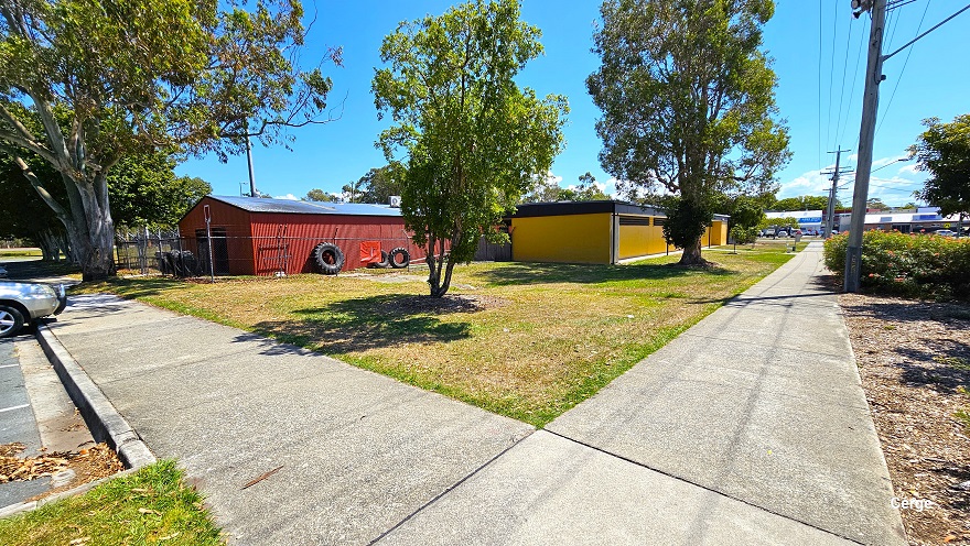 Intersecting concrete pathway with a rough-looking surface when walking from the left side of the main entrance of the facility where the dedicated carpark is located. There is a wide grass bank and multiple tall trees that have wide canopy cover for shading before the mustard-yellow brick building. Close to the mustard yellow building is a red corrugated building on the left side, enclosed by a chain-linked fence. Placed against the walls are large and small black tires.