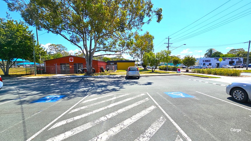 Dedicated carpark at Deception Bay Aquatic Centre. There are white diagonal line markings enclosed by a white rectangular marking between two accessible car park spaces. The accessible car park spaces can be distinguished by the blue background with a white accessible logo for persons with mobility disabilities. The red corrugated building, the mustard-yellow brick building, and the yellow sunshade sails can be seen from this position.