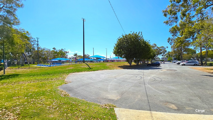 Curb section of the dedicated carpark at Deception Bay Swimming Pool. The tall trees with abundant foliage offer shade to several cars parked. The 50m, 8-lane pool with 3-tiered steps and the blue sun shade sails are visible from this position. The contoured grass banks that surround the aquatic facilities provide privacy from the surrounding roads.