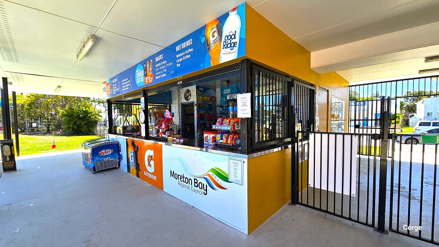 On the central entry is a floor-to-ceiling black gate on the left half, while on the right is a low black security pool gate that serves as a barrier entry to facilitate the ticket-paid entry into the mustard yellow brick administration building. On the right wall is a large yellow and white signboard for operation hours, entry fees, and the holidays of the facility. Next to it is a yellow door and a glass window for the entry tickets.
