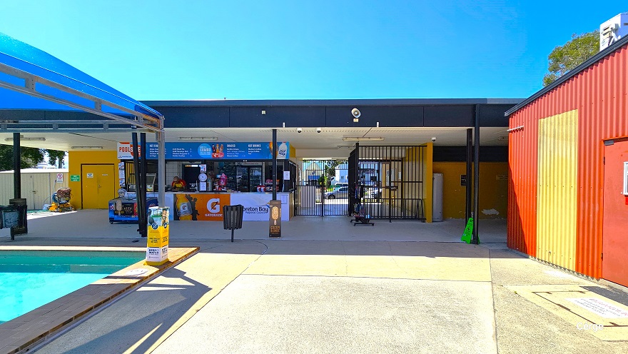 View of the central entry with black iron rod gates and the reception. On the left side of the reception with retail is a small space that leads to the male and female bathrooms, plus an accessible bathroom with a press-button entry. The Cérge app logo holding a QR code is placed behind a blue fridge. A small section of the outdoor pool covered with blue sunshade sails and the red corrugated with yellow accents can be seen on the opposite sides.