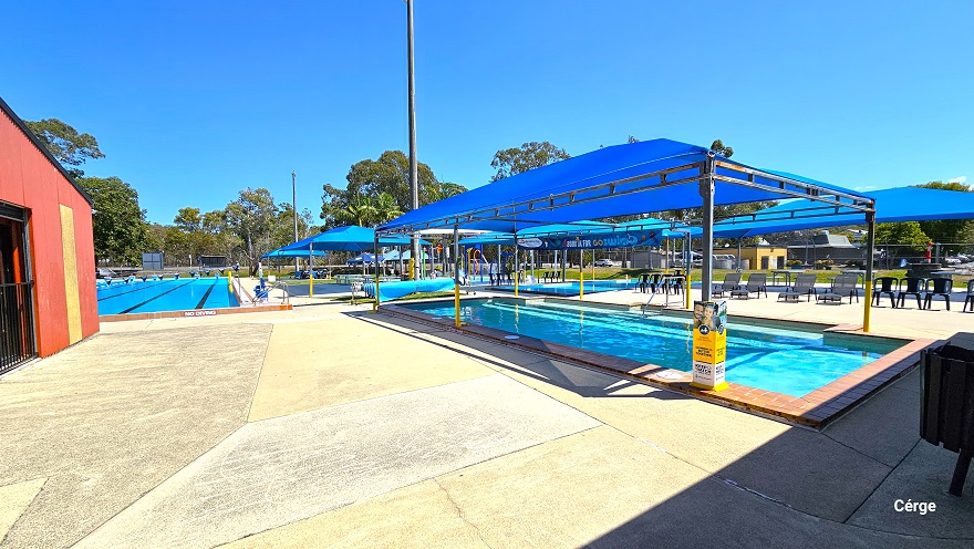 Outdoor aquatic facilities at Deception Bay Swimming Pool. There is ample step-free access right after the reception area and on the heath learn to swim pool. The pool is covered by a blue sunshade sail that is supported by grey and yellow metal frames. It has a brown brick pool coping that outlines the entire pool. On the right side is a wide blue sunshade sail with black plastic waterproof chairs. Other pools and blue sun shades are visible from this position.