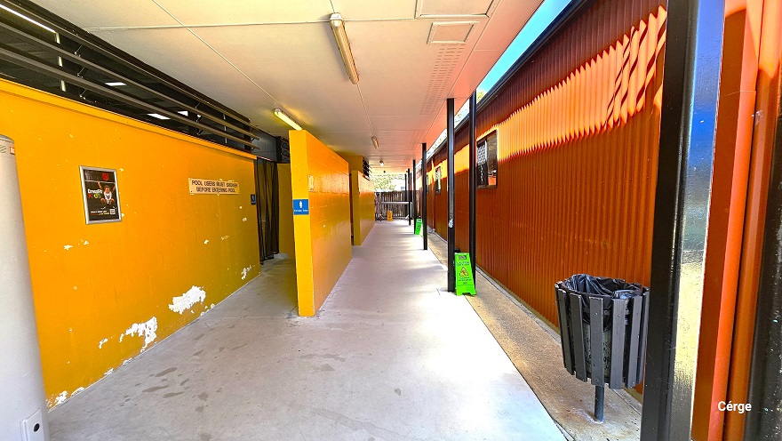Pathway leading to the bathrooms next to the red corrugated with black iron building. The bathroom area has a mustard yellow brick wall with small white patches. The entry point is obscured by another mustard yellow brick wall that separates it from the outside and another bathroom section. On the right side is a black wooden circular garbage bin with a black garbage bag and light green caution signs for wet floors.