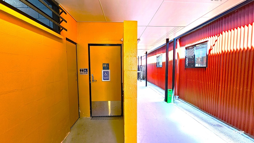 Accessible bathroom at Deception Bay Swimming Pool. On the bathroom door are small blue signs with a logo for persons with mobility disabilities. The lower part of the door is made of silver metal and it is outlined by black paint. The same sign can also be seen on the left wall of the door but on a silver sign. Another door is accessible on the left wall. This section can be found towards the end and is slightly obliterated by a small mustard-yellow brick wall.