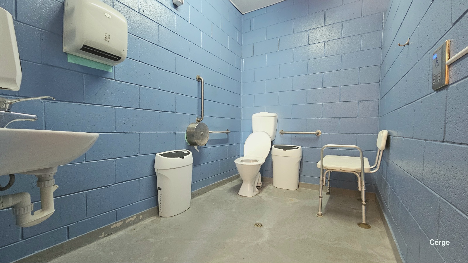 Inside view of the accessible bathroom at Deception Bay Swimming Pool. It has a light blue brick wall, white ceiling, and smooth grey floor. Found near the door are a wash basin, a liquid hand soap dispenser, and a white tissue paper dispenser mounted on the left wall. On the front and side of the toilet are two water softeners. Grab bars mounted on the walls and a white shower and bath seat are available to aid persons with mobility issues.