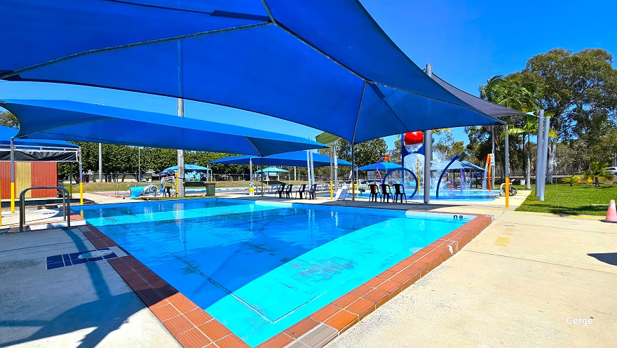 View of the heated learn-to-swim pool and shallow infant pool at Deception Bay Aquatic Centre. Next to the pools are blue sunshade sails with black waterproof plastic chairs and picnic tables. The sunshades have grey and yellow metal frame supports. All the pools have brown pool coping that outlines the pool shape. A white pool hoist is available next to the 50m, 8-lane outdoor Olympic lap pool.