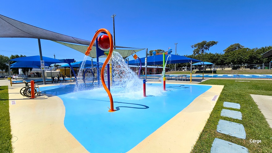 Children’s splash park with different water features and a giant water-dumping bucket. The splash park has a light blue floor and has zero depth. There is a white aquatic wheelchair with back support next to a red metal bollard. On the grass next to the splash park are grey squares with rough surfaces. Two triangular sunshades cover half of the splash park.