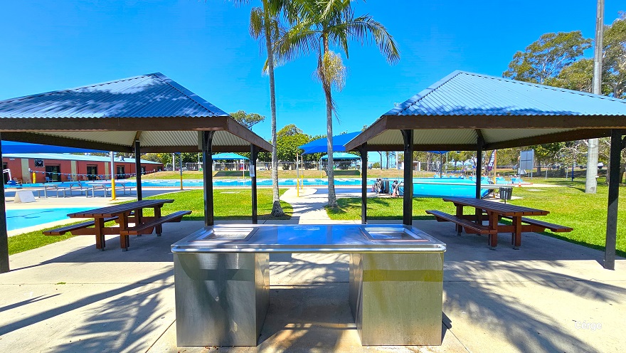 View in front of the stainless steel outdoor BBQ at Deception Bay Swimming Pool. Two park shelters with corrugated roofing with dark brown roof supports. Each shelter has a wooden picnic table. The outdoor BBQ is next to the splash park and coconut trees that partially shade the BBQ grilling station.