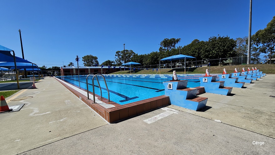 50m, 8-lane outdoor Olympic lap pool. On top of each of the blue 3-tiered stair steps numbered from 1 to 8 is an orange cone. The pool has a dark brown tile pool coping. Each pool lane is separated by blue, white, and red floating rope lanes. Strings of blue triangular flags overhang the pool. On the left side of the pool is a pool ladder with an inverted U handrail. The pool deck is a concreted floor with a rough surface.