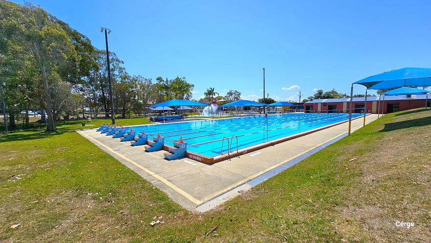 Outdoor 50m 8-lane pool, children’s splash water park, infants shallow pool, and heated learn-to-swim pool, as well as open grass areas for picnics and a large undercover BBQ at Deception Bay Aquatic Centre. The 50m, 8-lane pool has 3-tiered steps numbered from 1 to 8 for diving. On the left and right side of the pool are blue shade sails with seating for families. To the back is a rusty red-colored industrial building with yellow rectangular wall accents and a light blue roof.