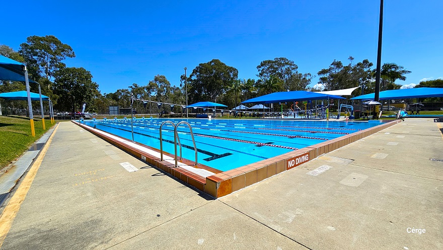 50m, 8-lane outdoor pool has 3-tiered blue steps numbered from 1 to 8 for diving. The pool deck has a noticeable yellow outline close to the border. On the left and right side of the pool are blue shade sails with seating for families. To the back is a rusty red-colored industrial building with yellow rectangular wall accents and a light blue roof. The tall trees offer substantial shading options and the open grass area around the outdoor aquatic area offers open space for an outdoor activity.