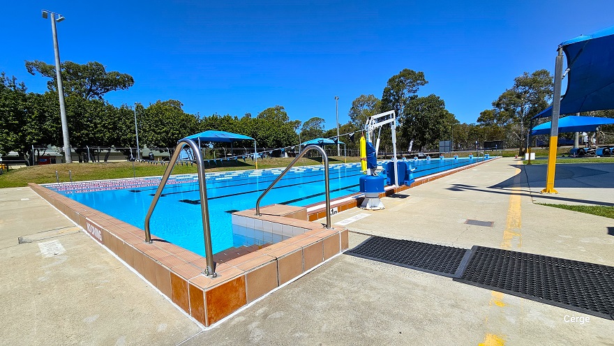 View of the stair access with steel handrails of the 50m, 8 lane Olympic outdoor pool. The image shows that the pool wall is white tile while the pool coping and stairs are a dark brown tile floor.  There is a small and large black mat close to the pool stairs. Closer to the pool stairs is a white pool hoist with blue seating to help move persons with mobility disabilities. A No Diving sign can be found on the side of the dark brown pool coping.