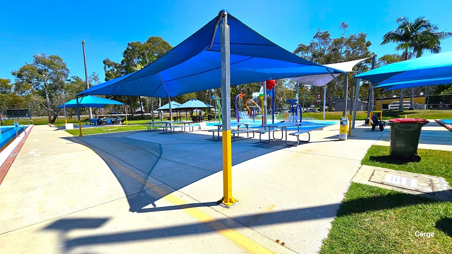 eating section between the 50m, 8 lane Olympic outdoor pool and the children’s splash park. The area is covered by a blue sunshade sail supported by grey and yellow metal frames. It has picnic tables, while to the right is a large grey garbage bin with wheels. The BBQ station, children’s splash park, and the heated learn-to-swim pool are visible from this position.