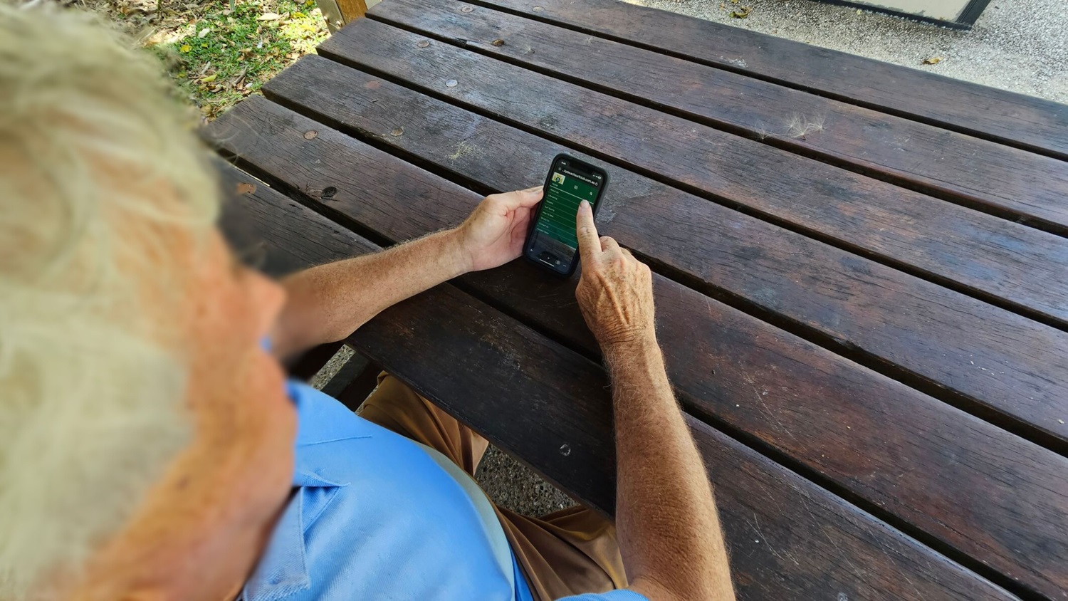 A person with light hair is sitting at a wooden picnic table, wearing a light blue shirt and beige trousers. They are holding a smartphone with both hands, using their right index finger to interact with the screen. The table has a weathered appearance with visible grain and some discolouration. In the background, there is grass and a small patch of concrete. A person with light hair is sitting at a wooden picnic table, wearing a light blue shirt and beige trousers. They are holding a smartphone with both hands, using their right index finger to interact with the screen. The table has a weathered appearance with visible grain and some discolouration. In the background, there is grass and a small patch of concrete.