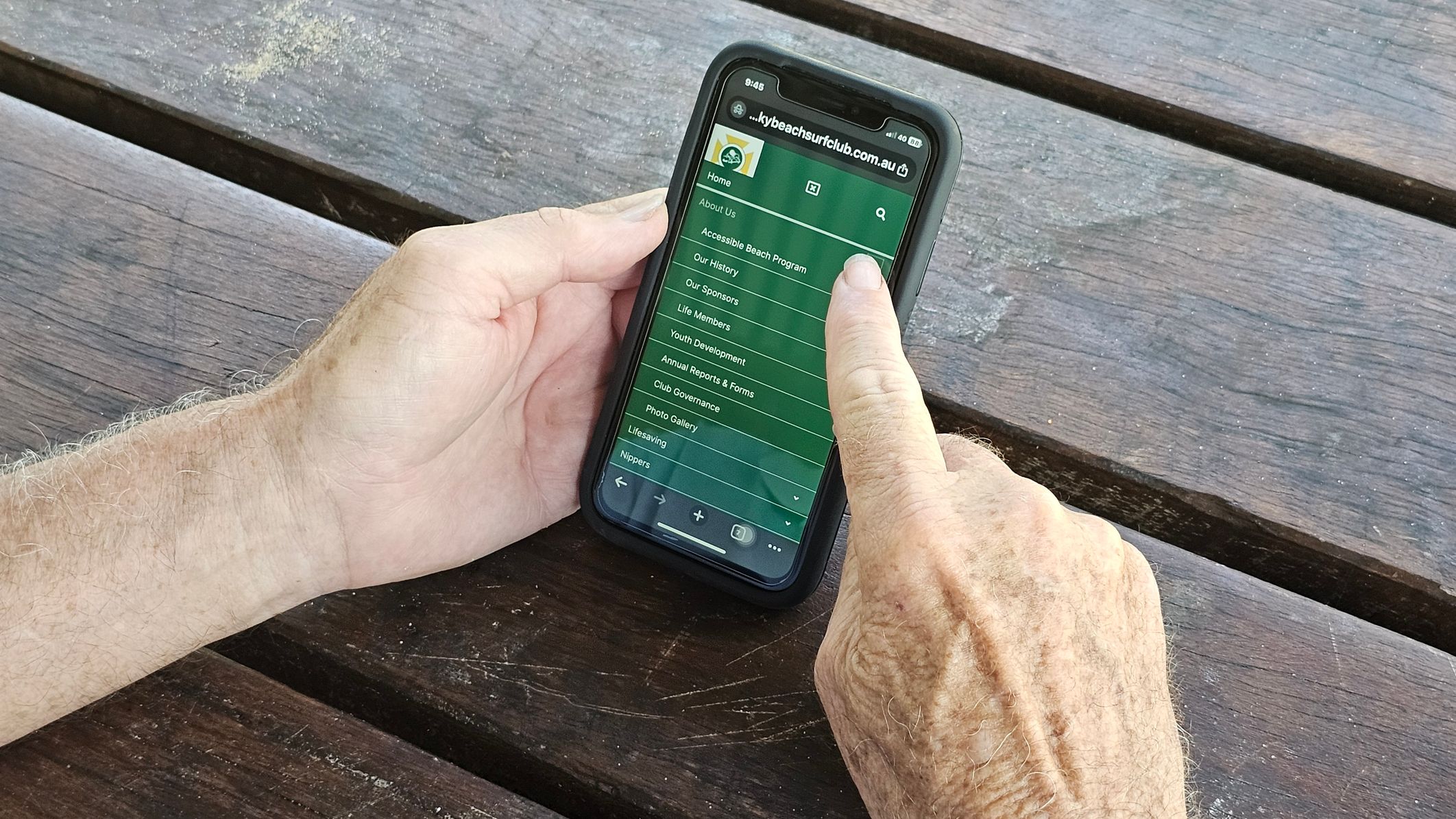 A person is holding a smartphone with both hands. The phone displays a website menu with options like "About Us" and "Our History." The background is a wooden surface with visible grain and some faded paint marks. The person's left hand supports the phone, while the right hand points at the screen. The phone has a black case. A person is holding a smartphone with both hands. The phone displays a website menu with options like "About Us" and "Our History." The background is a wooden surface with visible grain and some faded paint marks. The person's left hand supports the phone, while the right hand points at the screen. The phone has a black case.