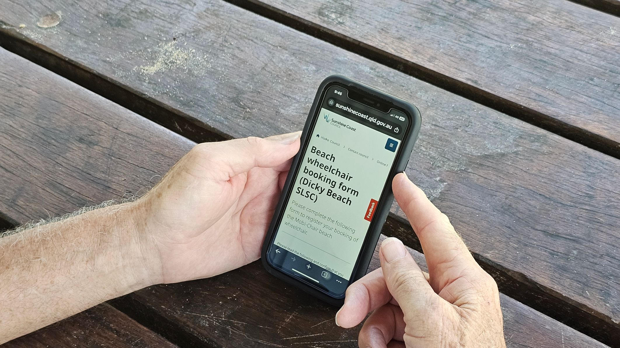 A person is holding a smartphone with both hands. The phone screen displays a website for booking a beach wheelchair at Dicky Beach SLSC. The background consists of a weathered wooden table with visible grain and some faded paint marks. The person's hands are positioned around the phone, with one thumb touching the screen. A person is holding a smartphone with both hands. The phone screen displays a website for booking a beach wheelchair at Dicky Beach SLSC. The background consists of a weathered wooden table with visible grain and some faded paint marks. The person's hands are positioned around the phone, with one thumb touching the screen.