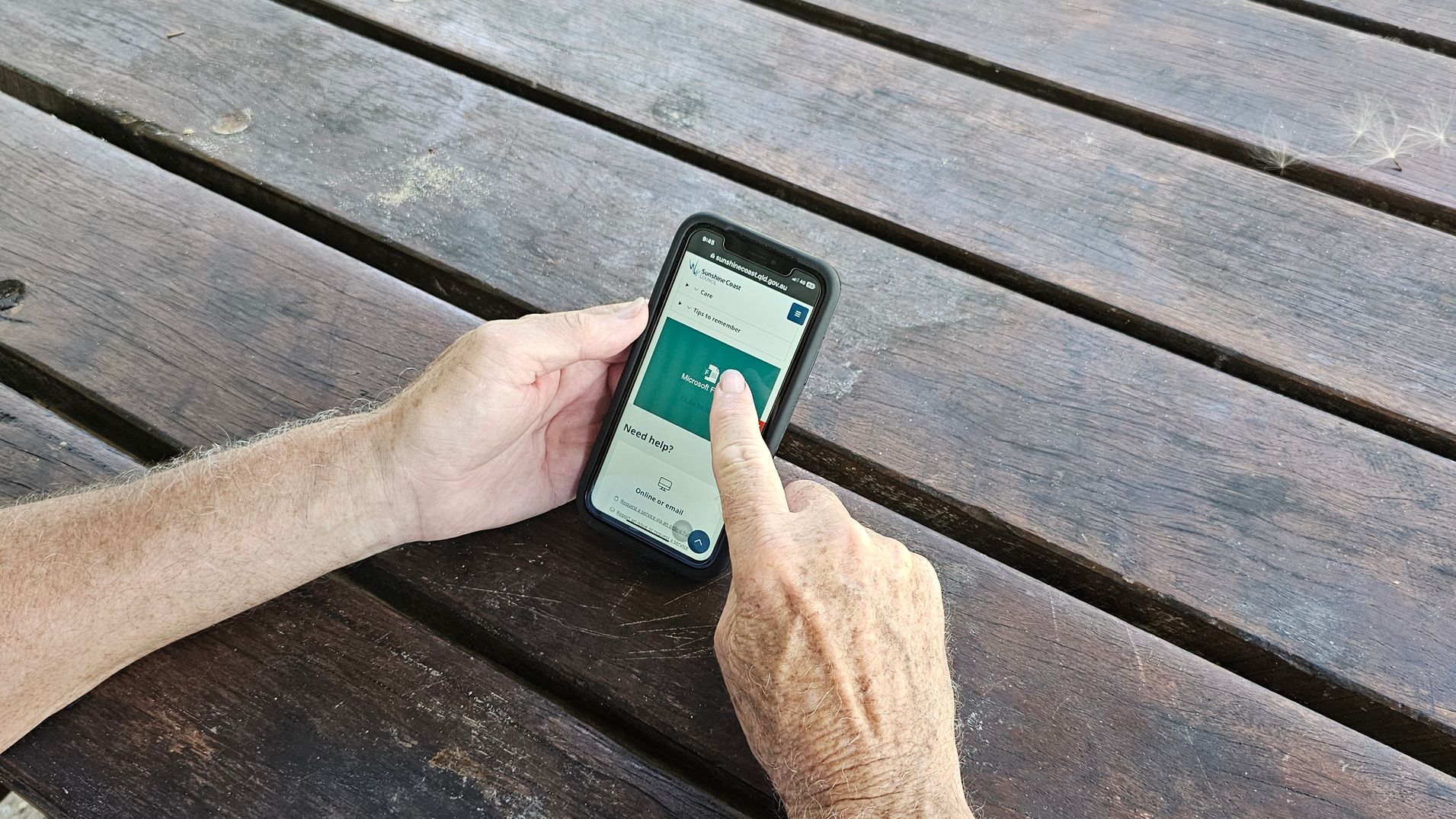 A pair of hands holding a smartphone with a black case, positioned over a weathered wooden table. The phone screen displays a website with a green section and text. The person's right index finger is touching the screen, interacting with the content. The table has visible grain and some discolouration, with gaps between the planks. A pair of hands holding a smartphone with a black case, positioned over a weathered wooden table. The phone screen displays a website with a green section and text. The person's right index finger is touching the screen, interacting with the content. The table has visible grain and some discolouration, with gaps between the planks.