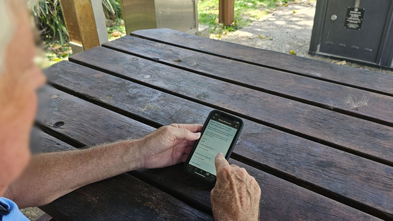 A person with light skin is sitting at a weathered wooden picnic table, holding a smartphone in both hands. The phone screen displays a webpage with text. In the background, there is greenery and a paved path. A black metal bin is visible to the right. The table has a few scattered seeds or fluff on its surface. A person with light skin is sitting at a weathered wooden picnic table, holding a smartphone in both hands. The phone screen displays a webpage with text. In the background, there is greenery and a paved path. A black metal bin is visible to the right. The table has a few scattered seeds or fluff on its surface.