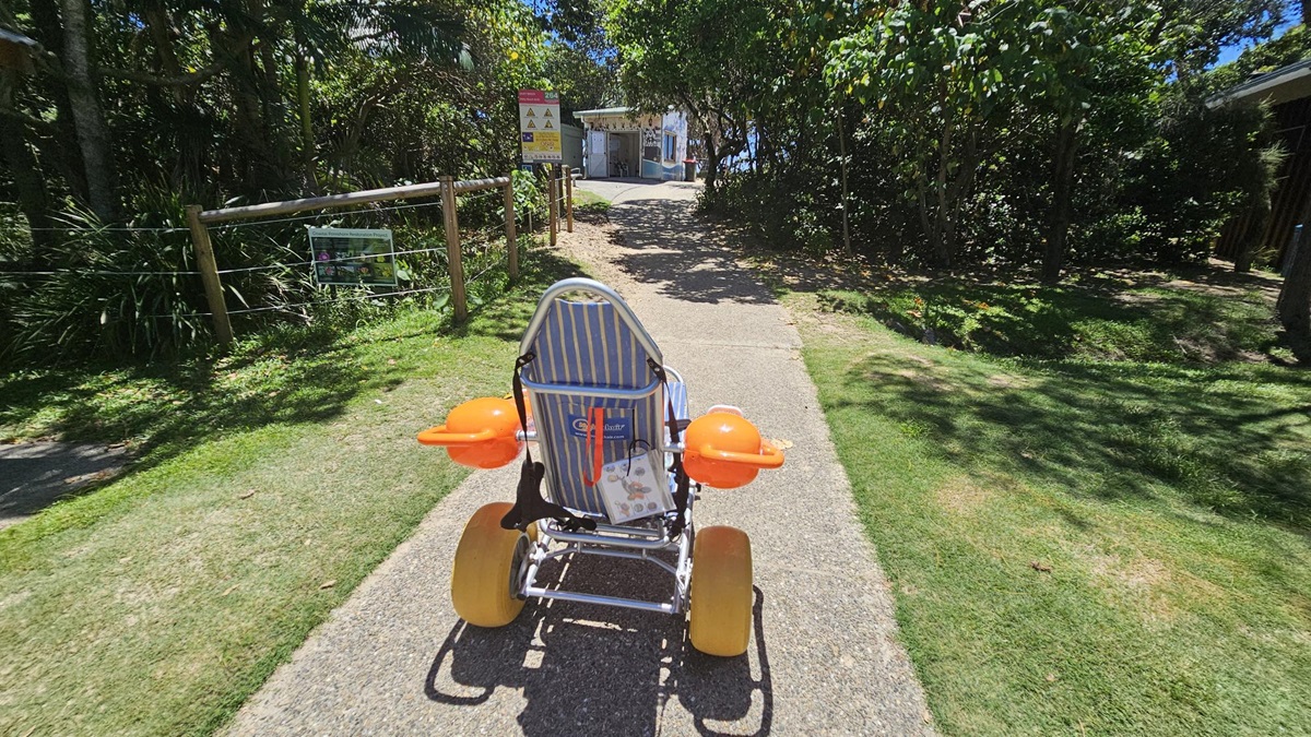 On the left, a wooden fence with wire runs alongside green bushes and trees, with a metal sign attached displaying information. On the right, grass and dense trees create shade over the area. In the middle, a path made of coarse concrete leads uphill towards a small white building with an open door and signs near it. In the foreground, a beach wheelchair with yellow wheels, orange armrests, and a blue-and-white striped backrest sits on the path. On the left, a wooden fence with wire runs alongside green bushes and trees, with a metal sign attached displaying information. On the right, grass and dense trees create shade over the area. In the middle, a path made of coarse concrete leads uphill towards a small white building with an open door and signs near it. In the foreground, a beach wheelchair with yellow wheels, orange armrests, and a blue-and-white striped backrest sits on the path.