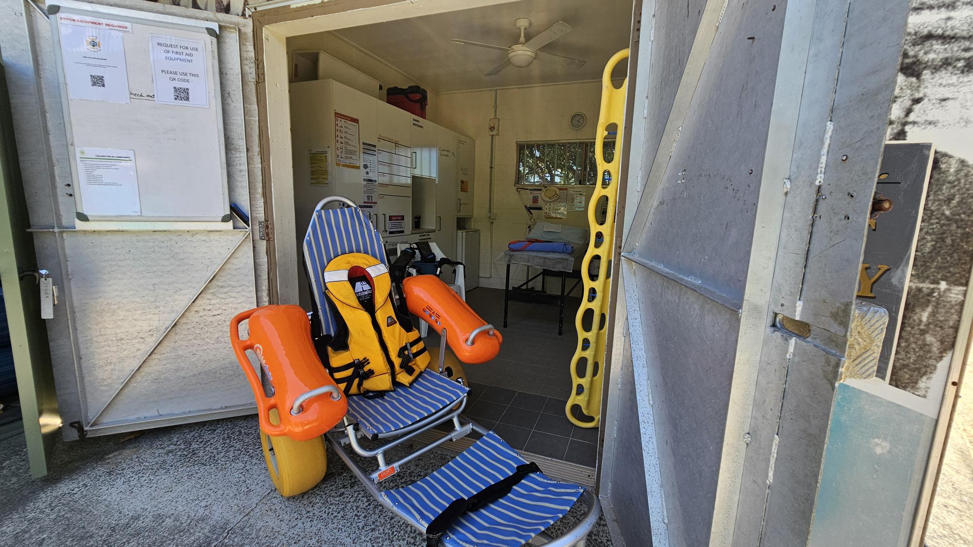 On the left, an open door with white panels features several notices and documents, including a sign with QR codes. On the right, a partially open metal door reveals a yellow spine board leaning against the frame. In the middle, a blue-and-white striped beach wheelchair with large yellow wheels and an orange life jacket is positioned on a textured concrete floor. The background contains a tiled floor, shelves with equipment, a table with folded towels, and a ceiling fan. On the left, an open door with white panels features several notices and documents, including a sign with QR codes. On the right, a partially open metal door reveals a yellow spine board leaning against the frame. In the middle, a blue-and-white striped beach wheelchair with large yellow wheels and an orange life jacket is positioned on a textured concrete floor. The background contains a tiled floor, shelves with equipment, a table with folded towels, and a ceiling fan.