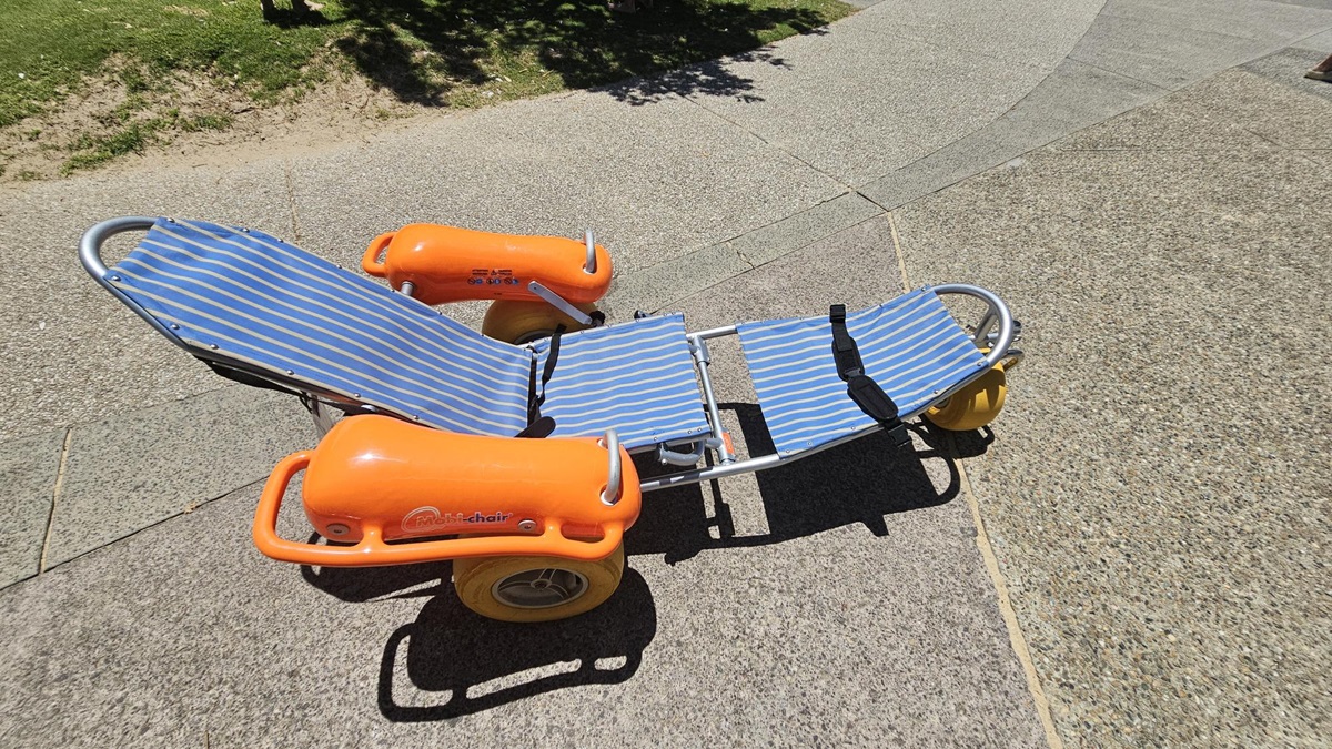 On the left, a green grassy area with uneven patches is visible. On the right, a textured walkway of light grey and beige paving stones extends diagonally. In the middle, an orange-and-blue striped beach wheelchair with yellow wheels is placed, its metal frame and inflatable armrests prominent. The foreground is covered with pebbled paving, and there are faint shadows from nearby trees cast on the ground. On the left, a green grassy area with uneven patches is visible. On the right, a textured walkway of light grey and beige paving stones extends diagonally. In the middle, an orange-and-blue striped beach wheelchair with yellow wheels is placed, its metal frame and inflatable armrests prominent. The foreground is covered with pebbled paving, and there are faint shadows from nearby trees cast on the ground.