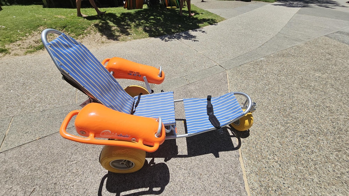 On the left, a small grassy area with uneven patches merges into a curved paved pathway. On the right, the pathway continues with a textured mix of grey and beige tones. In the middle, a beach wheelchair with blue-and-white striped seating, bright orange armrests, and large yellow wheels rests on the ground. The foreground features a pebble-textured surface, and faint tree shadows fall across the scene. On the left, a small grassy area with uneven patches merges into a curved paved pathway. On the right, the pathway continues with a textured mix of grey and beige tones. In the middle, a beach wheelchair with blue-and-white striped seating, bright orange armrests, and large yellow wheels rests on the ground. The foreground features a pebble-textured surface, and faint tree shadows fall across the scene.