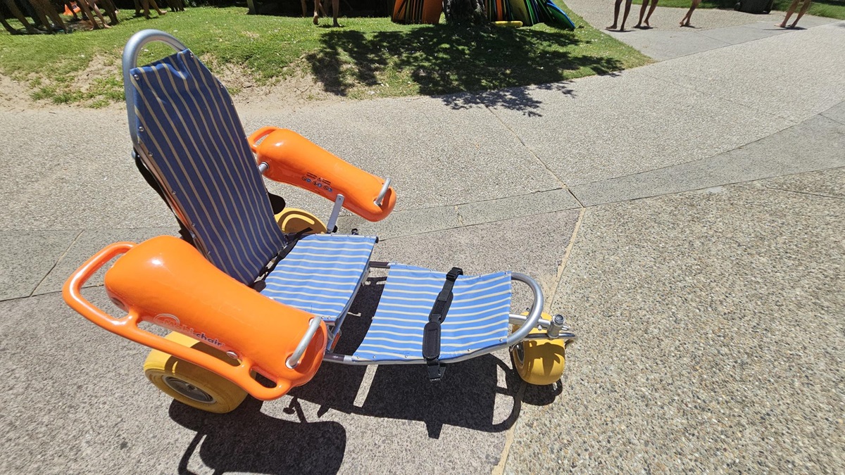 On the left, a small grassy area with uneven patches merges into a curved paved pathway. On the right, the pathway continues with a textured mix of grey and beige tones. In the middle, a beach wheelchair with blue-and-white striped seating, bright orange armrests, and large yellow wheels rests on the ground. The foreground features a pebble-textured surface, and faint tree shadows fall across the scene. On the left, a small grassy area with uneven patches merges into a curved paved pathway. On the right, the pathway continues with a textured mix of grey and beige tones. In the middle, a beach wheelchair with blue-and-white striped seating, bright orange armrests, and large yellow wheels rests on the ground. The foreground features a pebble-textured surface, and faint tree shadows fall across the scene.