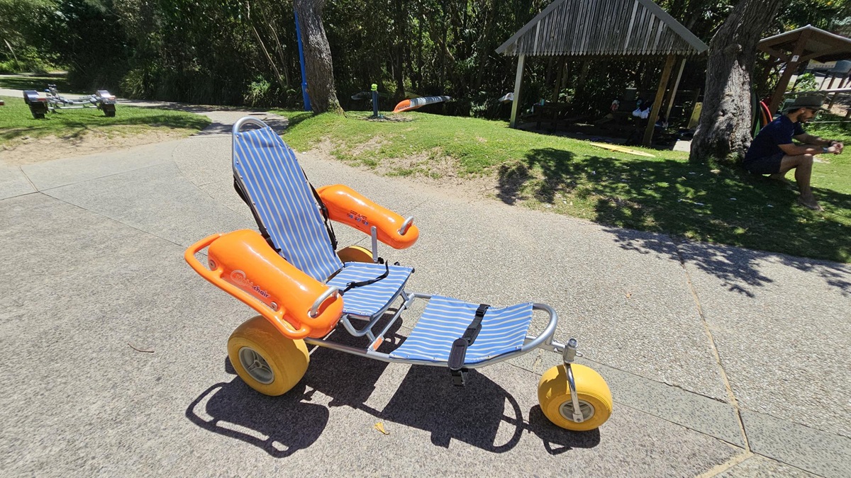 On the left, a green grassy area with a small pathway merging into a paved section. On the right, a patch of grass with a man sitting under a wooden shelter supported by poles alongside a tree trunk. In the middle, a beach wheelchair has a blue-and-white striped seat, orange armrests, and large yellow wheels. The floor consists of light grey textured pavements with faint shadows from nearby trees. There is no visible ceiling. On the left, a green grassy area with a small pathway merging into a paved section. On the right, a patch of grass with a man sitting under a wooden shelter supported by poles alongside a tree trunk. In the middle, a beach wheelchair has a blue-and-white striped seat, orange armrests, and large yellow wheels. The floor consists of light grey textured pavements with faint shadows from nearby trees. There is no visible ceiling.